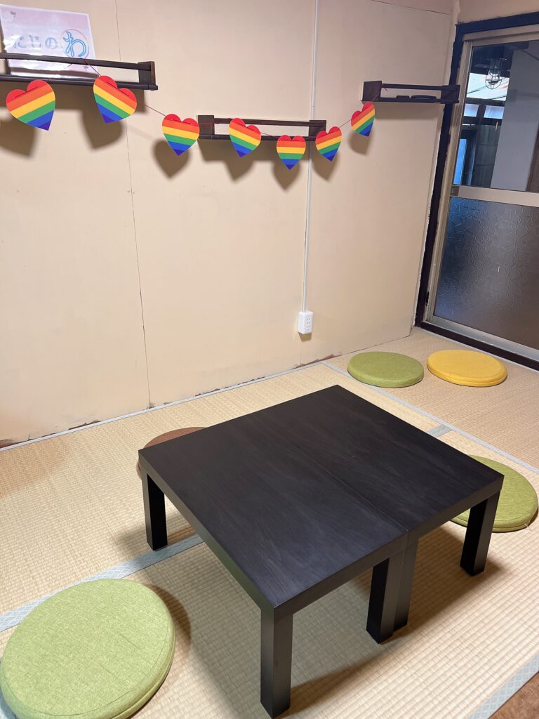 中の様子。畳の部屋に黒いテーブルと、円い生緑と黄色の座布団が並んでいる。壁にはハート型のプライドフラッグがかかっている。
