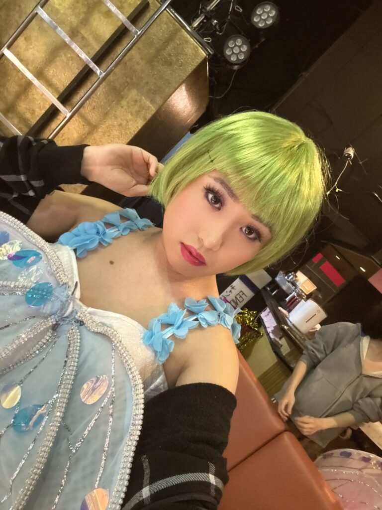 あかるい黄緑色のボブウィッグをかぶり、スパンコールがあしらわれた水色のドレスを着て自撮りをする、華やかなステージメイクの虎鉄さん。