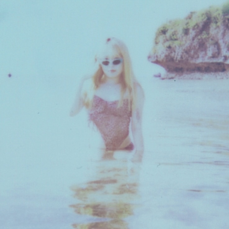 サングラスをかけ、ピンク色の水着を着て海の中に佇む、金髪の中里虎鉄さん。写真全体が淡い水色がかっている。