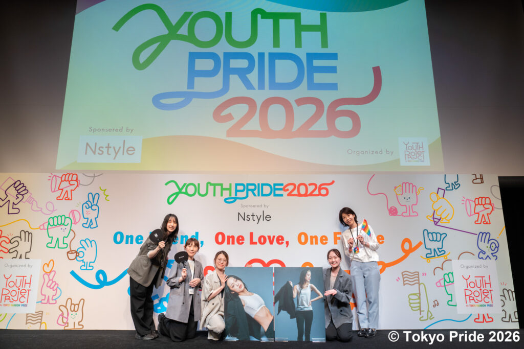「YOUTH PRIDE 2026」の巨大スクリーンの前で、写真パネルやレインボーフラッグを持って笑顔で記念撮影をする5人のNstyle社員の様子。