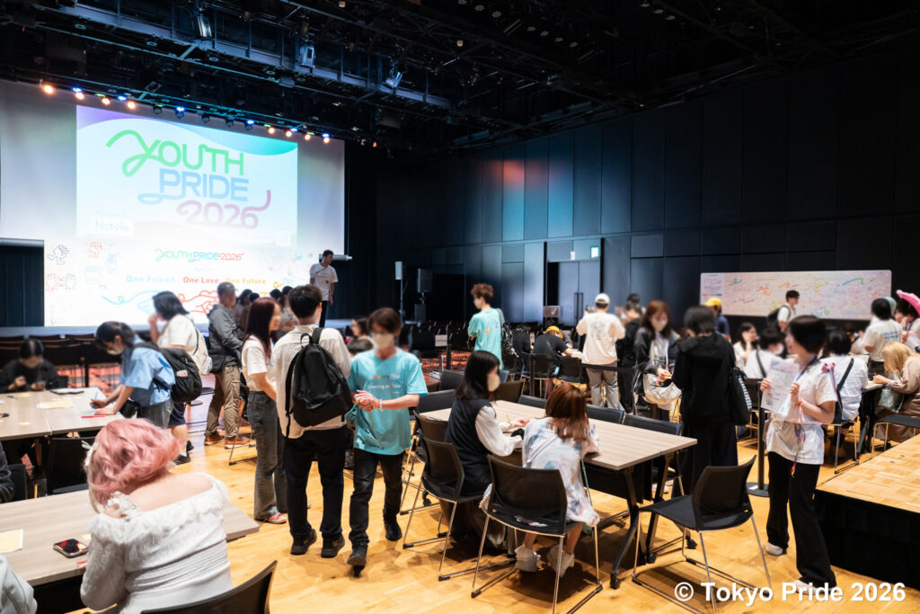 スクリーンに「YOUTH PRIDE 2026」のロゴが映し出されたイベント会場で、交流したりテーブルに着席したりする参加者たちの様子。右下には© Tokyo Pride 2026の著作権表記。